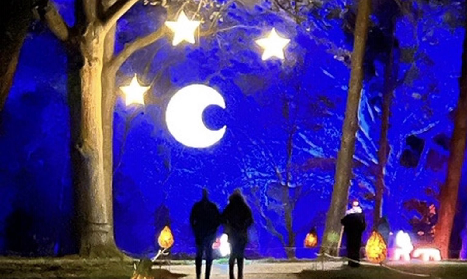 Two people stand between trees hung with bright stars and a present moon