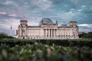 The colonnaded Bundestag with domed roof set against a cloud-filled sky with greenery in foreground.