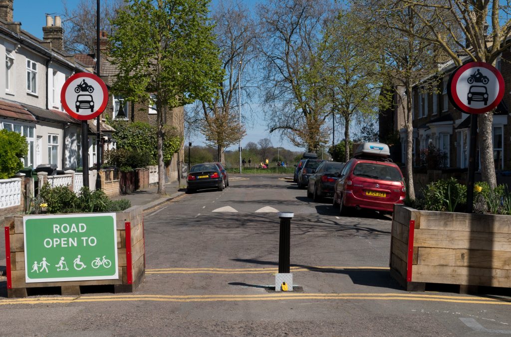 A small quiet tree-lined road in Oxford - part of a Low Traffic Neighbourhood. There are parked cars outside some of the houses. But the road is closed to cars at one end, preventing through traffic. The white on green sign has the words ROAD OPEN TO ... followed by small graphic images representing children playing, a child on their scooter, a person ion a wheelchair and a bicycle.