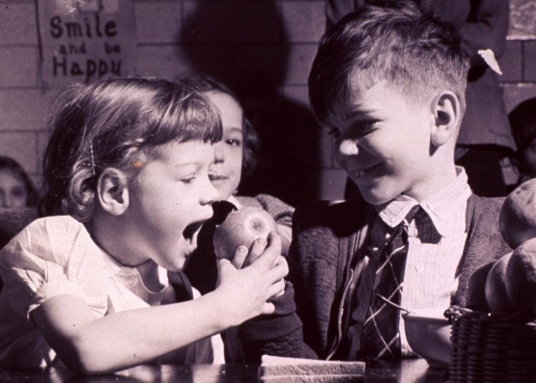 Children in a school cafeteria: girl about to bite an apple, 1950s. National Library of Medicine.