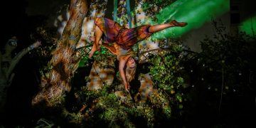 A female trapeze artist suspended upside down from a rope in a woodland