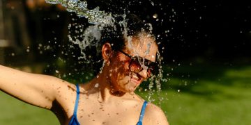 Woman in a blue swimsuit and sunglasses gets drenched by a wide stream of water outdoors, smiling as droplets spray around her.
