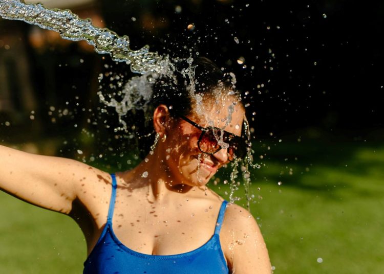 Woman in a blue swimsuit and sunglasses gets drenched by a wide stream of water outdoors, smiling as droplets spray around her.