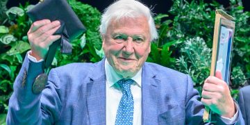 Older man in a light blue suit raises a dark folder in one hand and a framed document in the other, smiling outdoors.