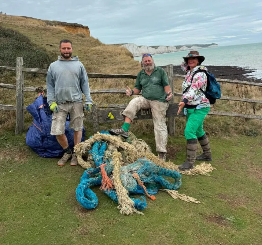 Three volunteers pose by a wooden fence on a coastal hillside with a tangled pile of blue and beige ropes, sea cliffs in the background.