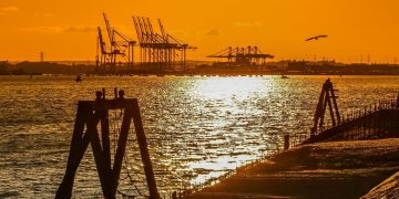 Silhouetted harbor at sunset with crane silhouettes in the distance and a wooden pier in the foreground.