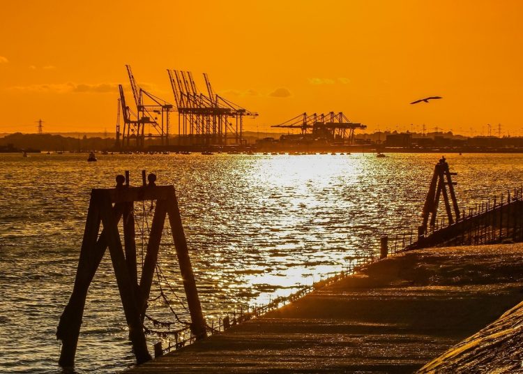 Silhouetted harbor at sunset with crane silhouettes in the distance and a wooden pier in the foreground.