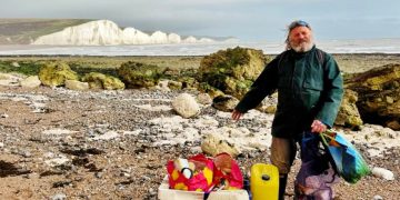Man wearing dark jacket on a pebbly beach, sorting and collecting trash into boxes and bags with white cliffs in the background.