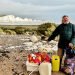 Man wearing dark jacket on a pebbly beach, sorting and collecting trash into boxes and bags with white cliffs in the background.