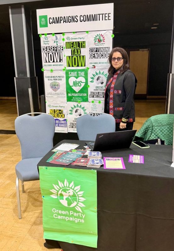 Woman with glasses standing beside a Green Party campaigns table; poster board behind shows slogans like 'Ceasefire Now' and 'Save the NHS.'