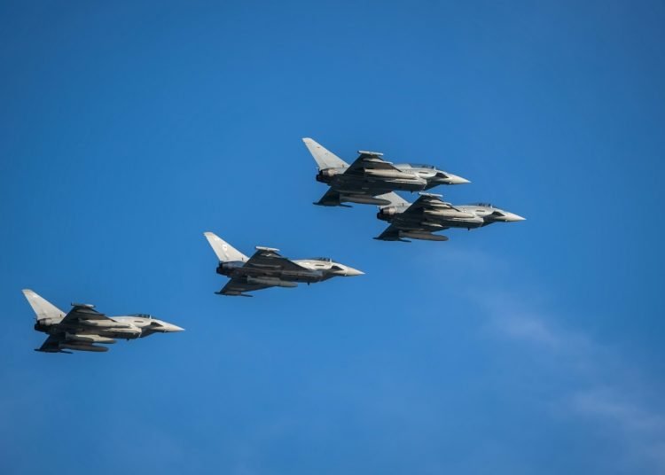 Four gray fighter jets fly in tight formation across a clear blue sky.