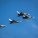 Four gray fighter jets fly in tight formation across a clear blue sky.