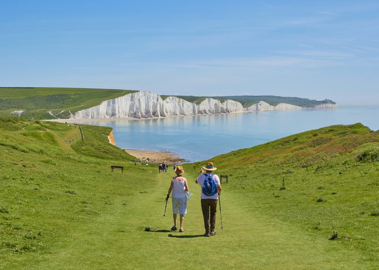 Hikers walking along a grassy cliff path toward a white chalk coastline and blue sea.