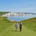 Hikers walking along a grassy cliff path toward a white chalk coastline and blue sea.