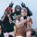 Group photo scene: photographer and a man with a phone in front of a blue wall, with a gorilla mascot behind them and several onlookers nearby.