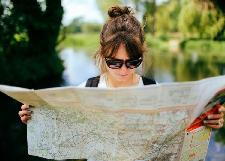 Woman with a bun and sunglasses holds and studies a large folded map outdoors in a green park area by water.