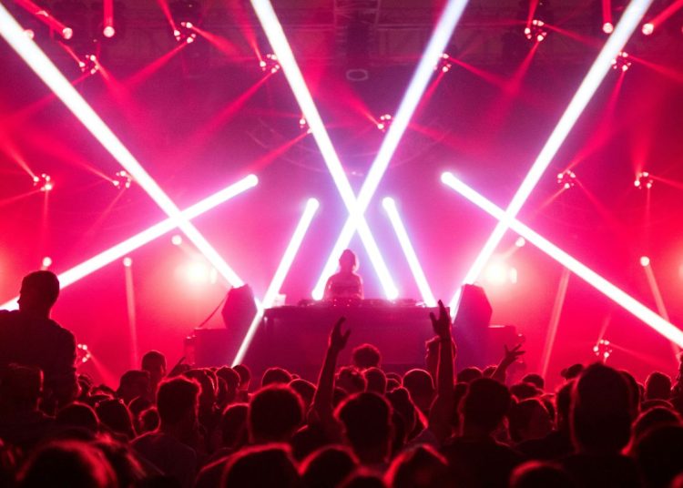 DJ on stage under red and pink laser lights, crowd raising hands at a high-energy concert.