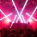 DJ on stage under red and pink laser lights, crowd raising hands at a high-energy concert.